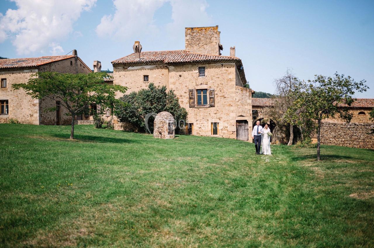 Un couple marche dans un jardin verdoyant devant un bâtiment en pierre ancienne sous un ciel partiellement nuageux.