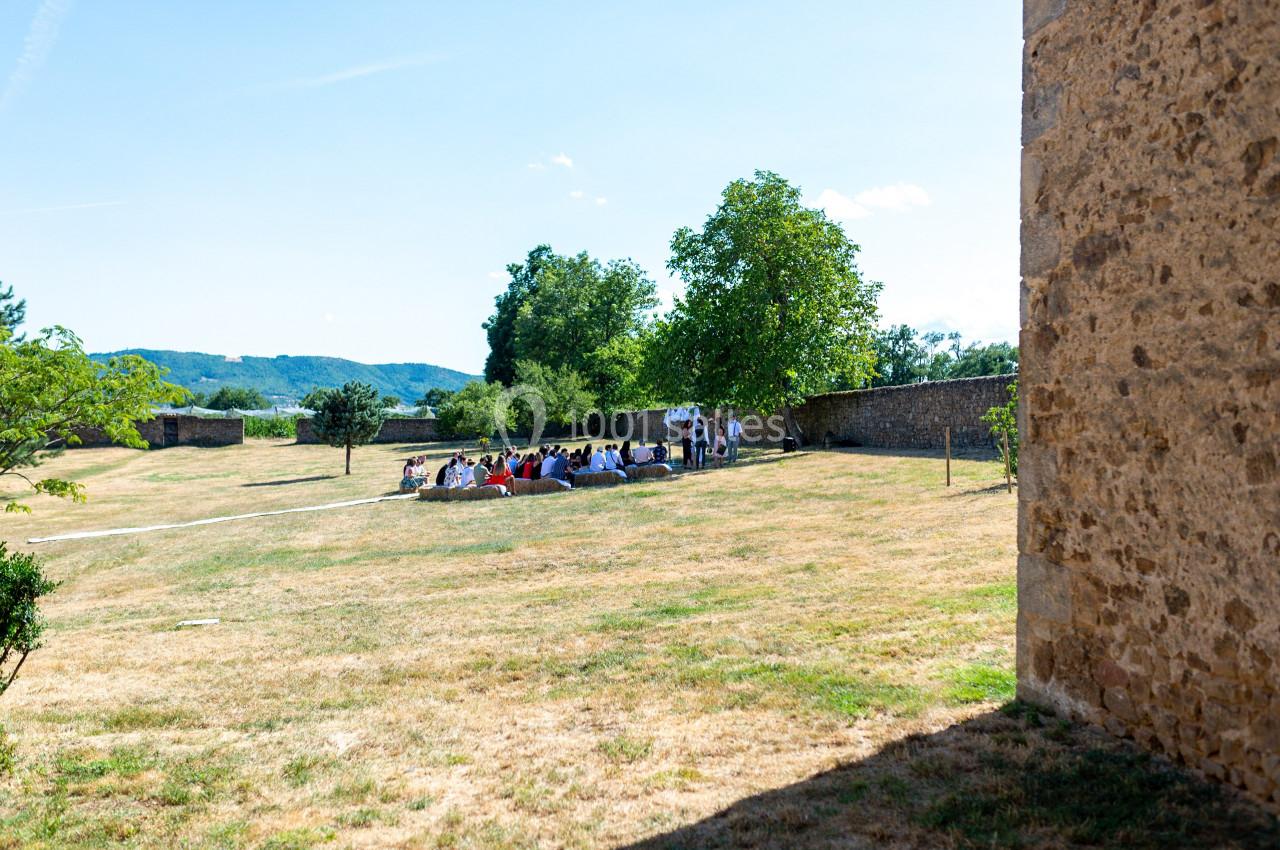 Un groupe de personnes assises en extérieur, sous un arbre, dans un espace herbeux près d'un mur en pierre.
