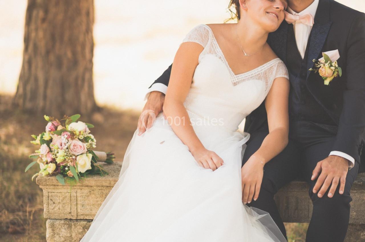 Un couple en tenue de mariage assis sur un banc en pierre, entouré de végétation et d'un bouquet de fleurs posé à côté.