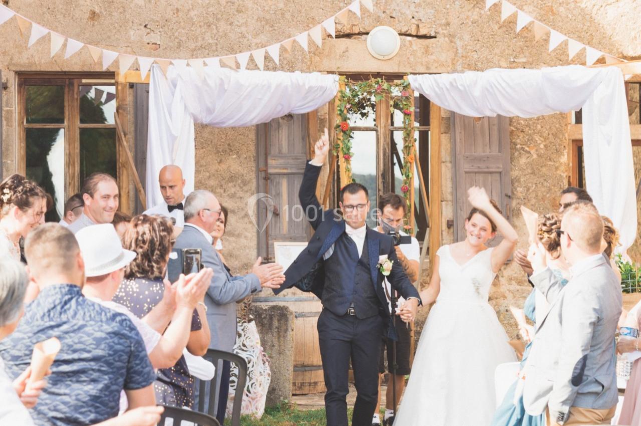 Un couple de mariés souriants sort sous les applaudissements des invités devant une bâtisse en pierre décorée.