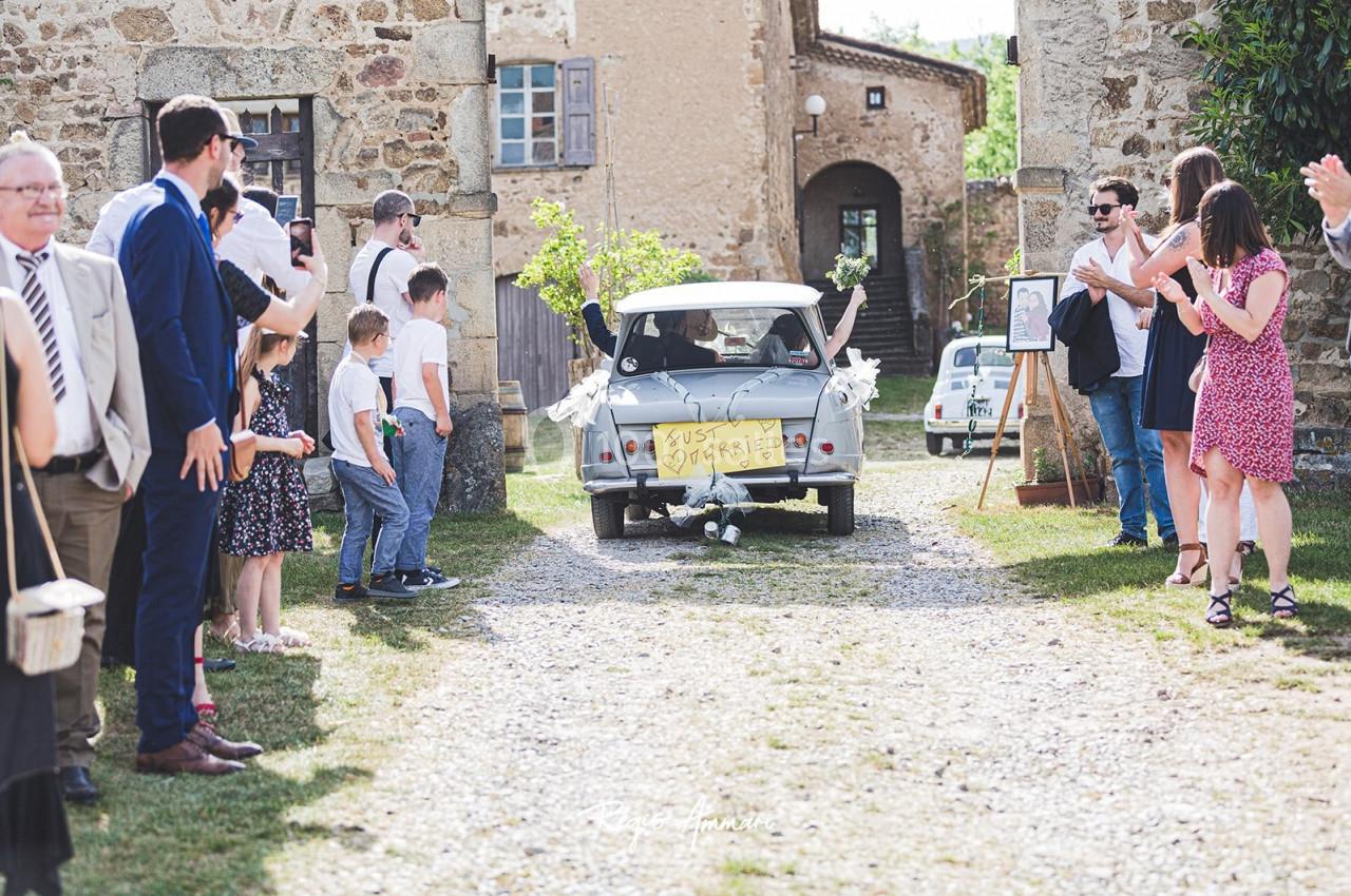 Une voiture décorée pour un mariage quitte un lieu entourée d'invités applaudissant.