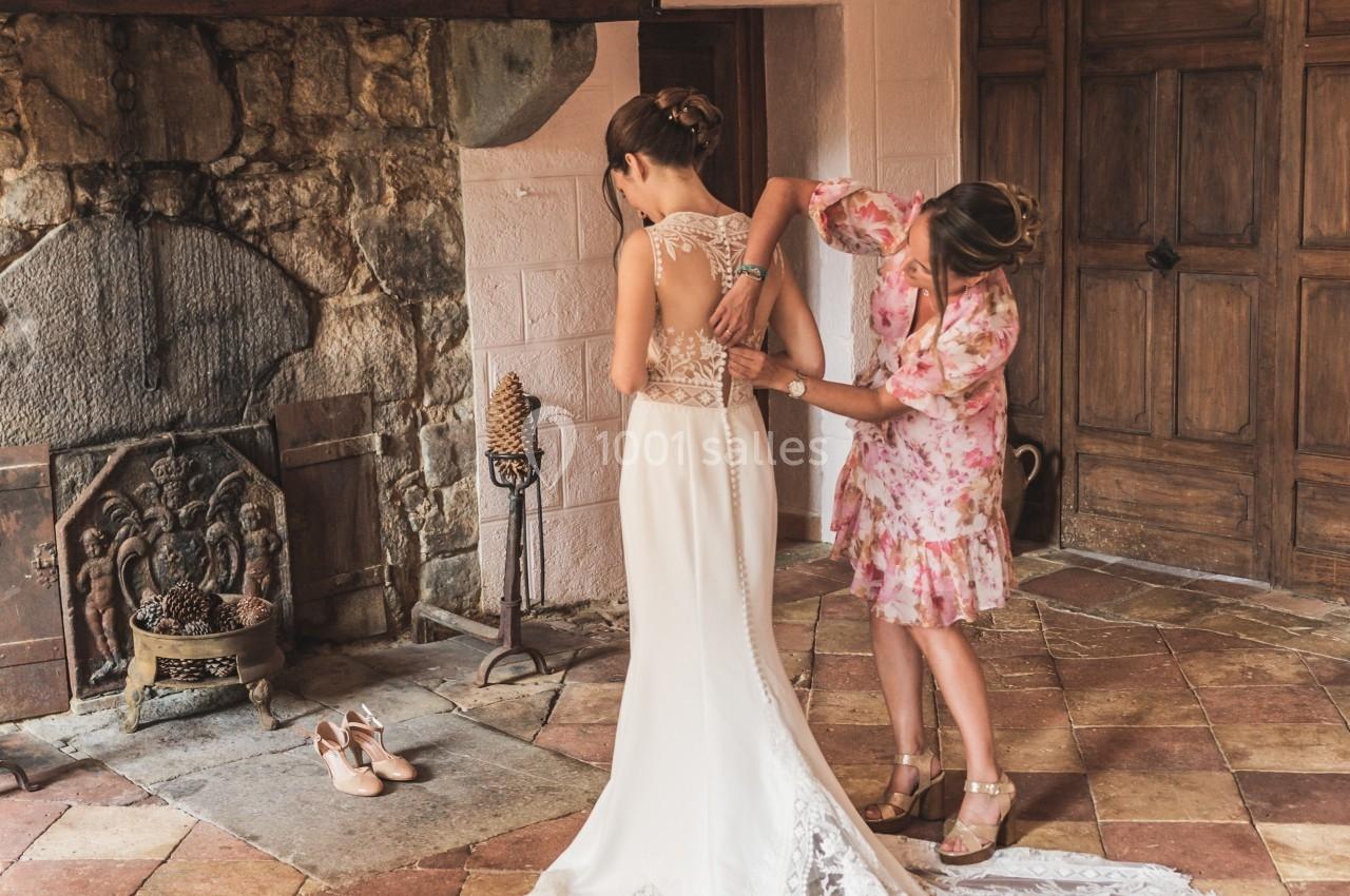 Une femme ajuste la robe de mariée d'une autre femme dans une pièce au sol en carreaux et cheminée en pierre.