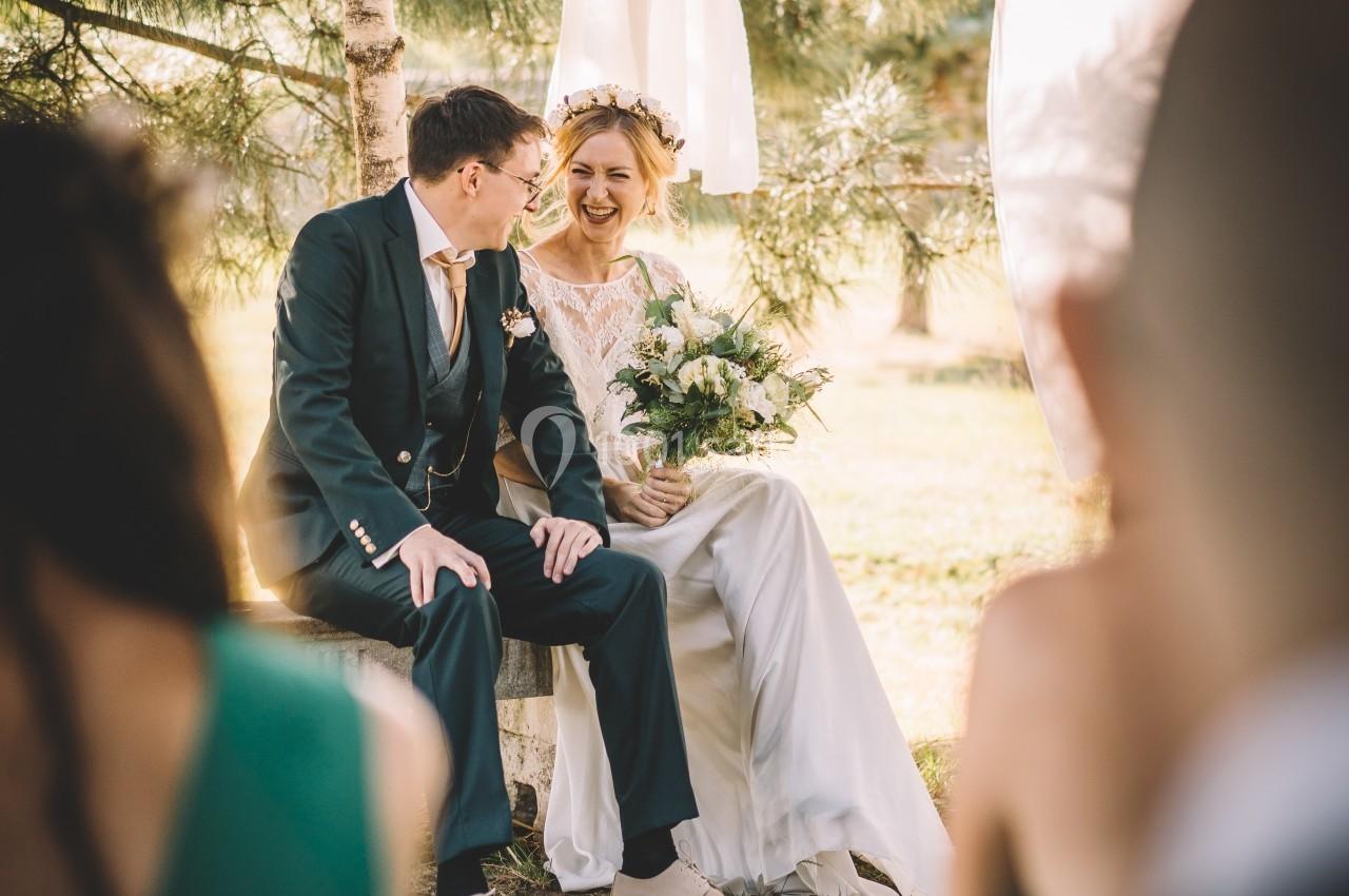 Un couple assis sur un banc en extérieur lors d'une cérémonie, entouré de verdure et de décorations en tissu blanc.