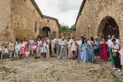 Groupe de personnes rassemblées dans une cour en pierre, certaines tenant des feuilles, ambiance festive en extérieur.