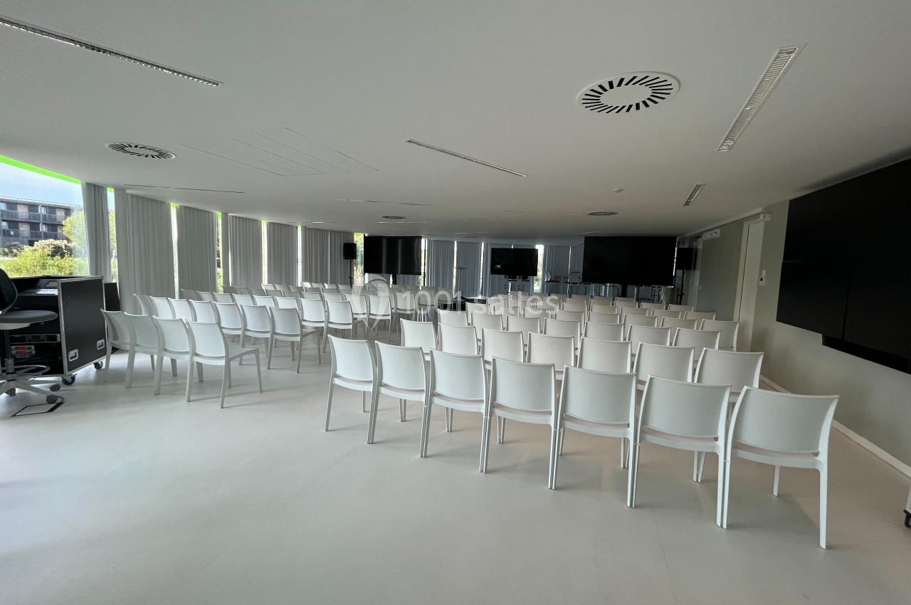 Salle de conférence lumineuse avec des rangées de chaises blanches alignées face à plusieurs écrans et fenêtres.