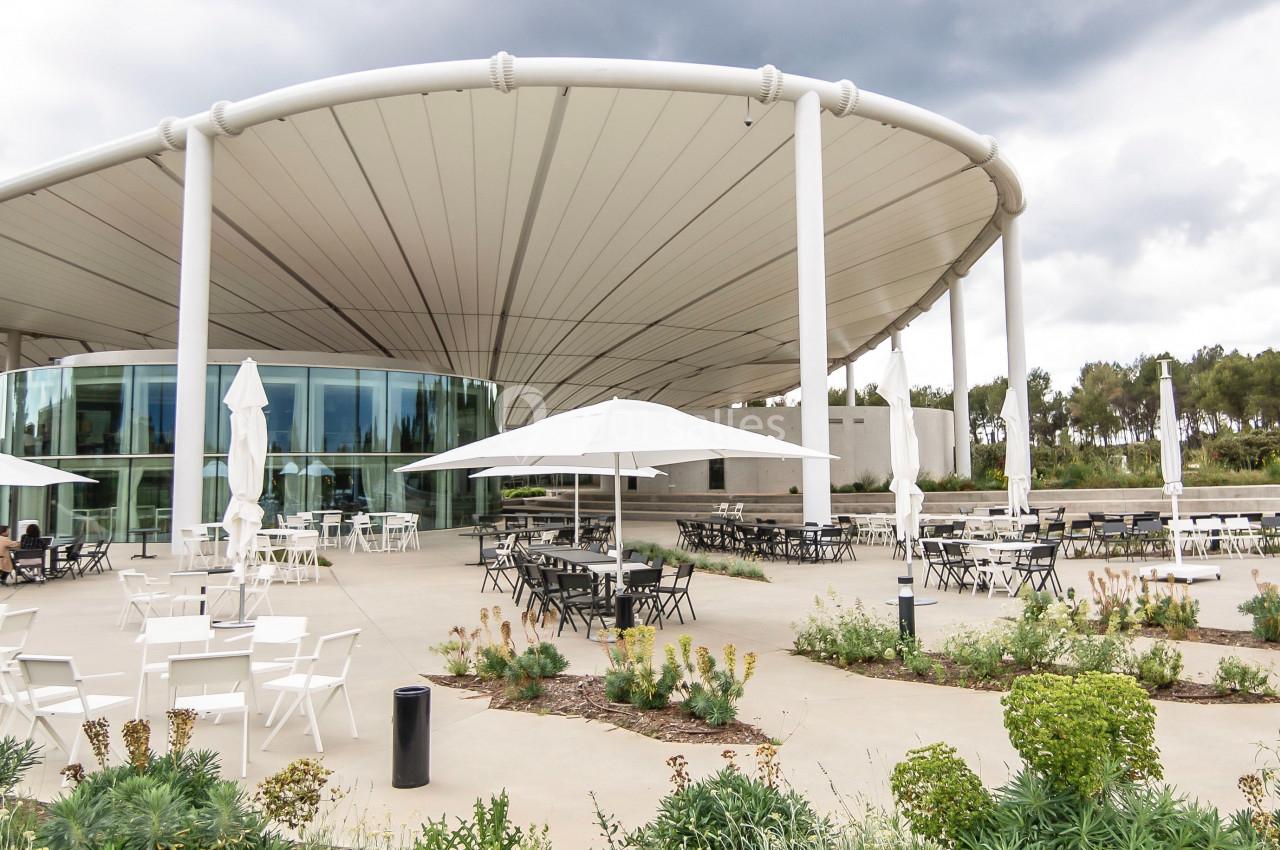 Terrasse extérieure avec tables, chaises et parasols devant un bâtiment moderne à toit arrondi sous un ciel nuageux.