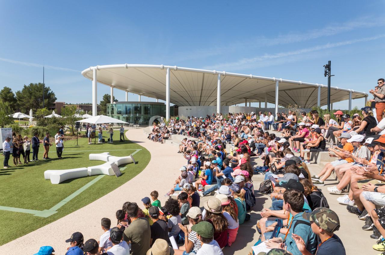 Foule assise en plein air devant une scène, sous un ciel dégagé, près d'un bâtiment moderne avec toit blanc.