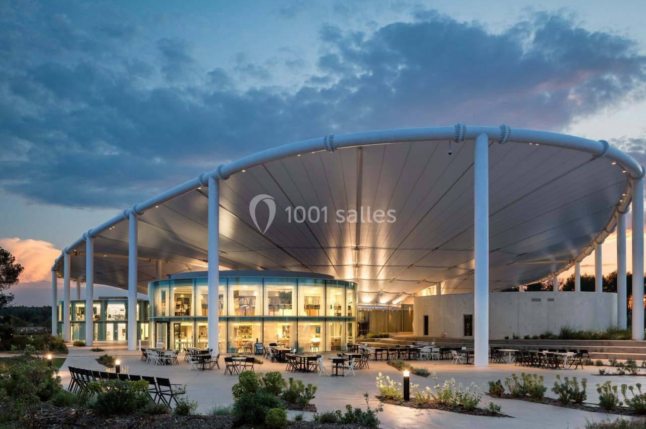 Bâtiment moderne avec une structure en toit incurvé, entouré de végétation et éclairé au crépuscule.