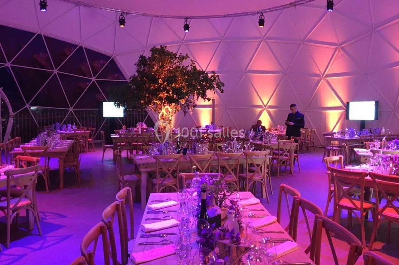 Salle de réception décorée avec des tables dressées, éclairage chaleureux et un arbre central illuminé.