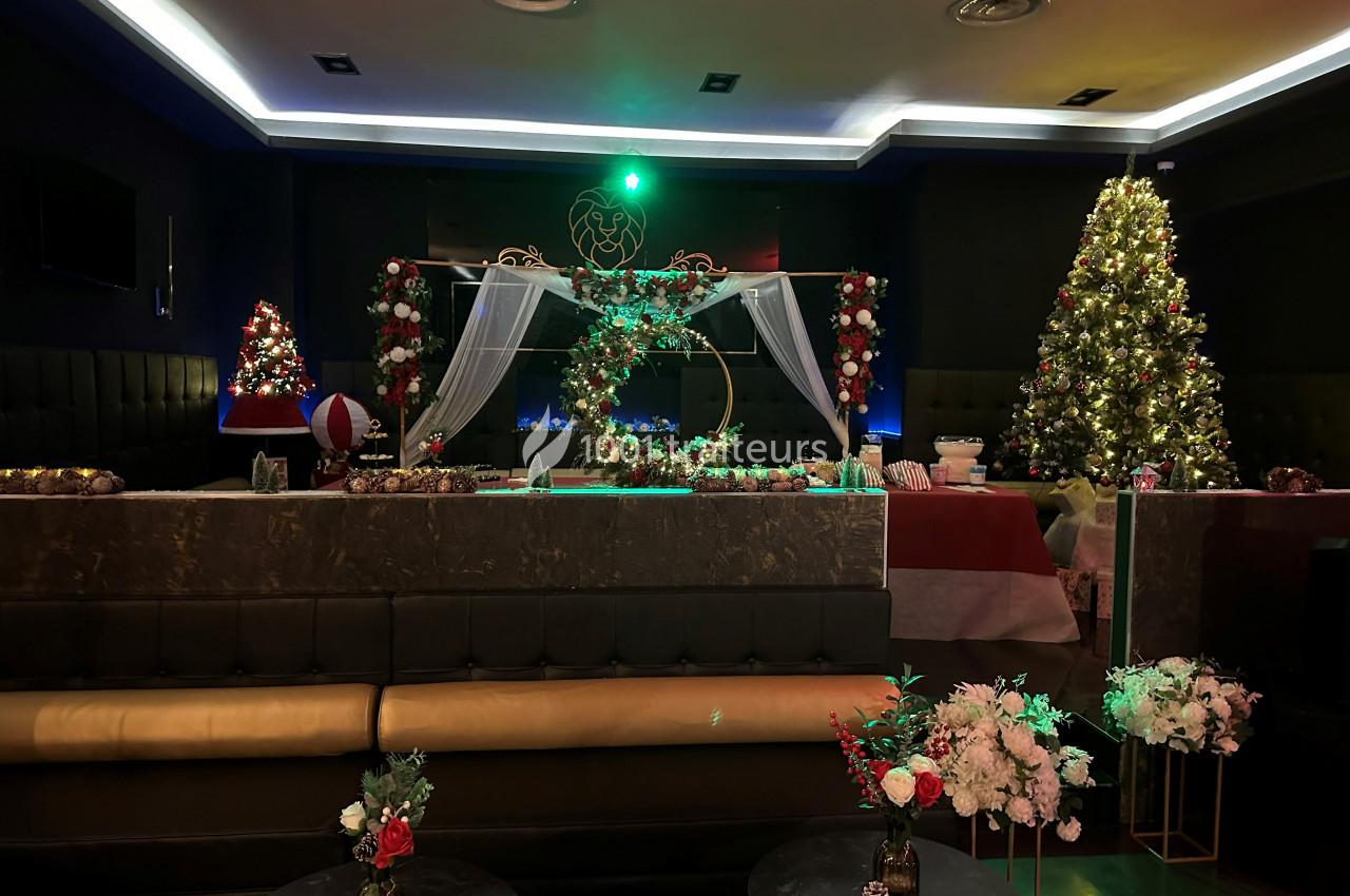 Salle décorée pour Noël avec sapins illuminés, guirlandes, fleurs et tables ornées dans une ambiance festive.
