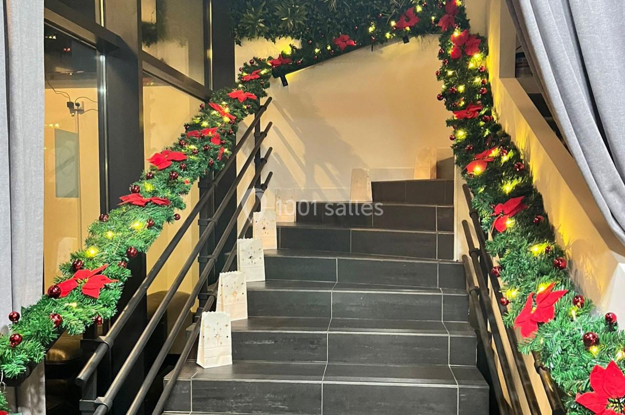 Escalier décoré de guirlandes lumineuses, de feuillage vert et de nœuds rouges, avec des sacs cadeaux posés sur les marches.