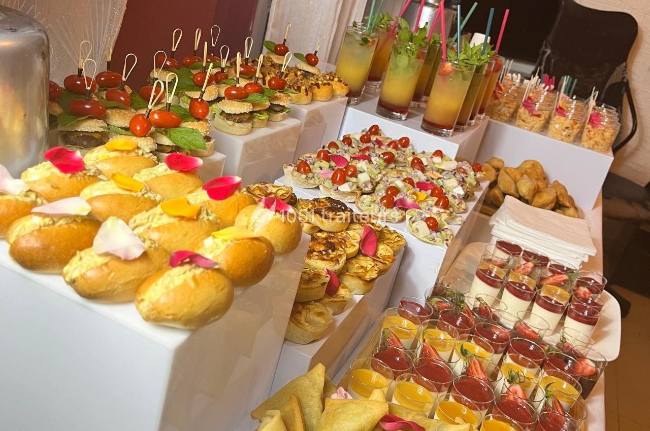 Buffet varié avec amuse-bouches, verrines, mini-sandwichs, desserts et boissons colorées sur des présentoirs blancs.