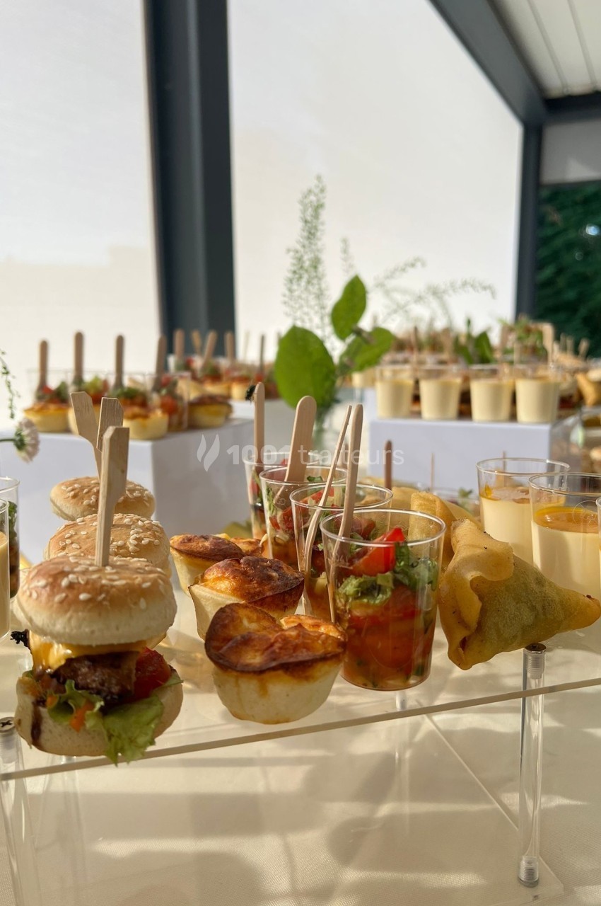 Buffet de verrines, mini-burgers, samoussas et bouchées salées disposés sur une table lumineuse en extérieur.