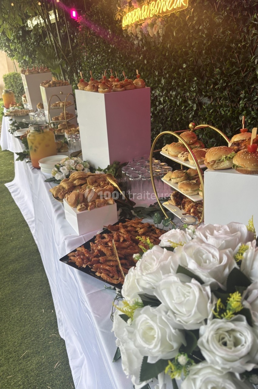 Buffet en extérieur avec divers plats, boissons, fleurs blanches en premier plan et décor végétal en arrière-plan.