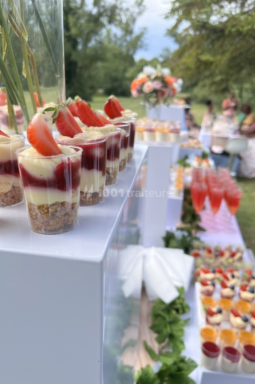 Desserts en verrines avec fraises et crème disposés sur une table en extérieur, entourés d'autres mets et boissons.