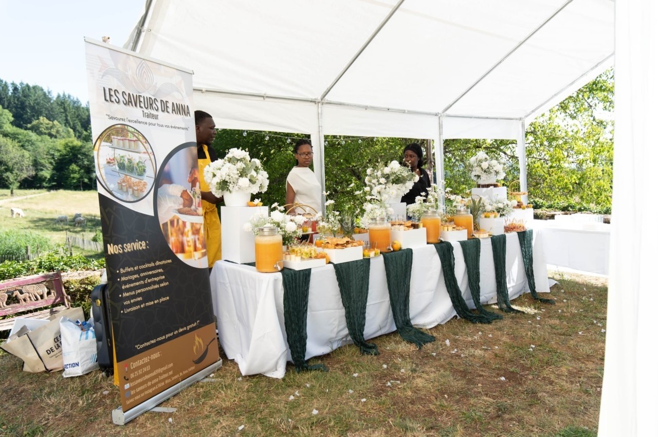 Stand de buffet sous une tente blanche, décoré de fleurs et boissons, avec trois personnes en arrière-plan.