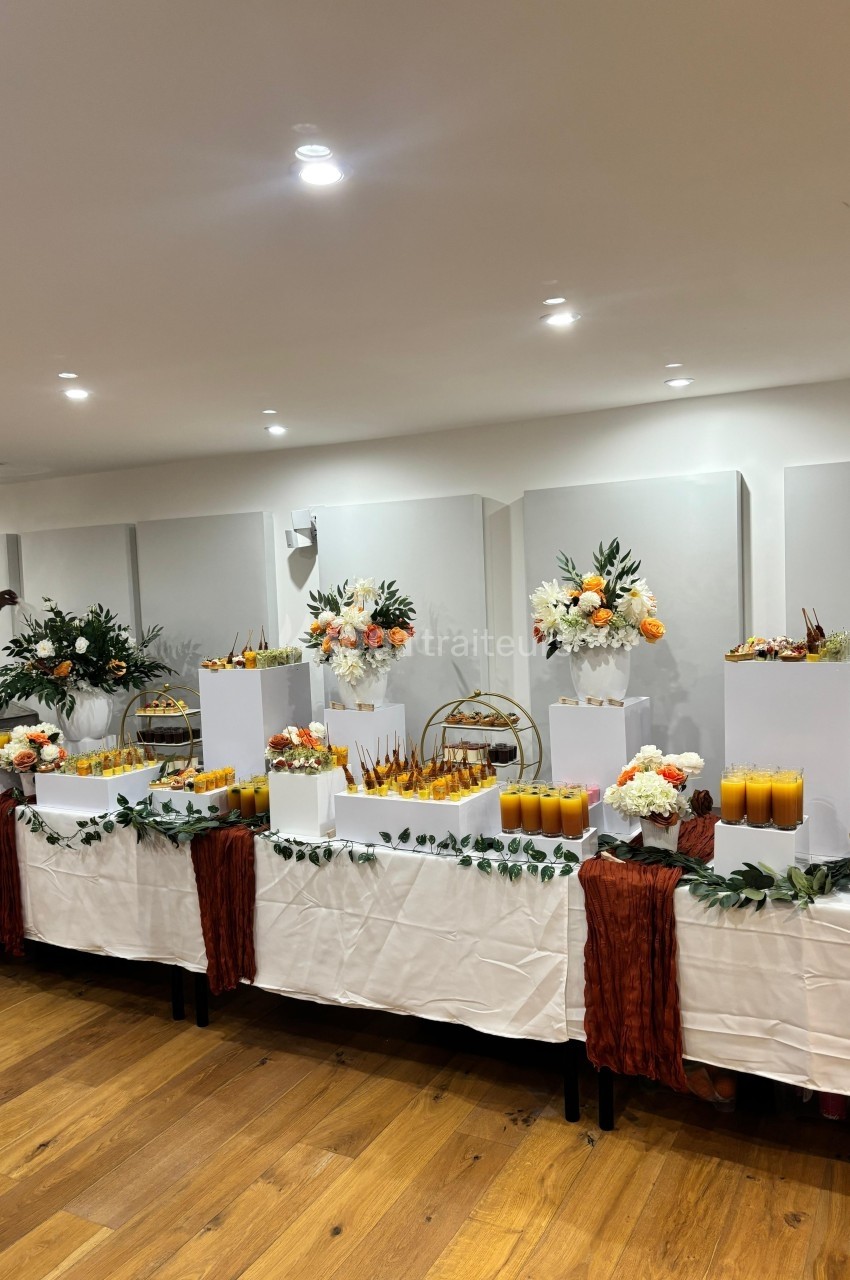 Table décorée pour un buffet avec fleurs, jus d'orange, verrines et éléments décoratifs sur nappes blanches.