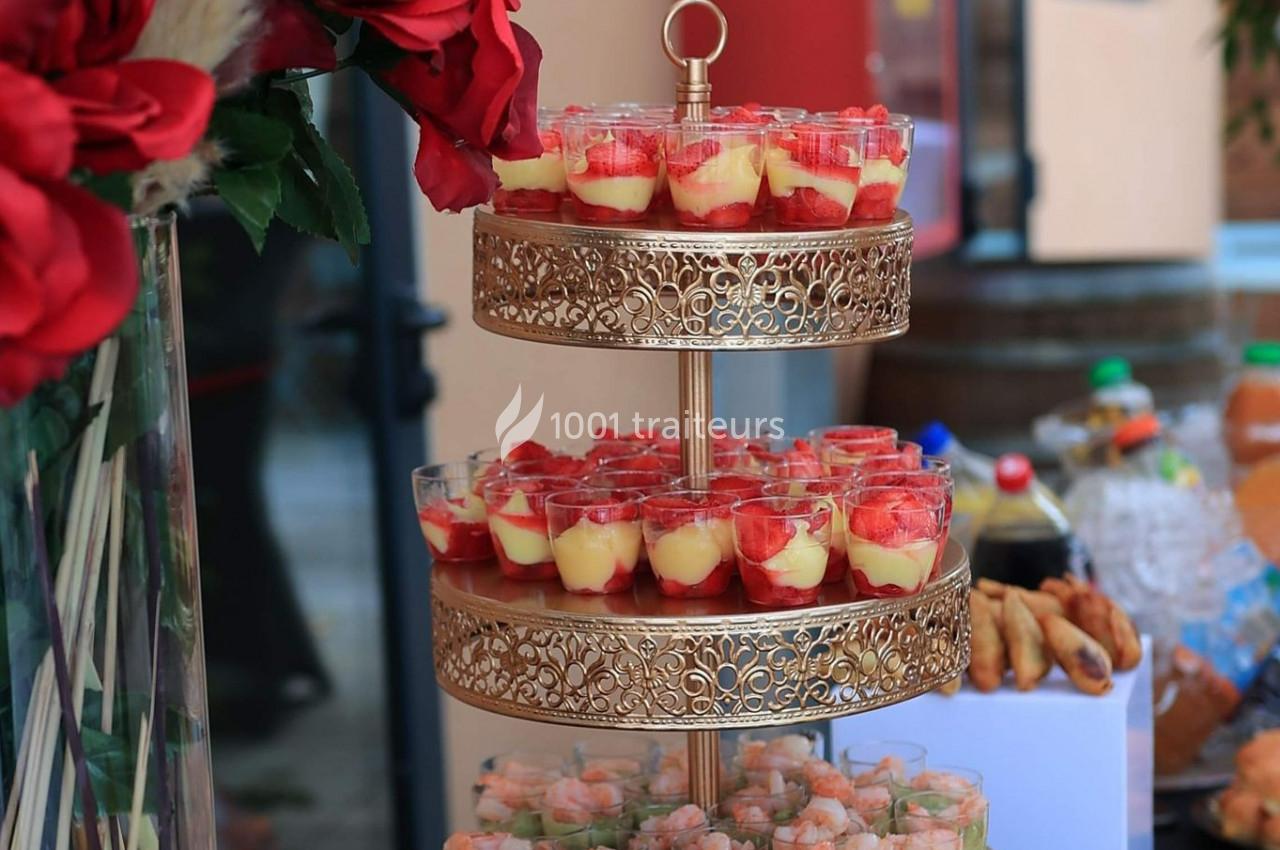 Plateau à deux étages en métal doré avec verrines de desserts aux fraises et crème, entouré de fleurs et boissons.