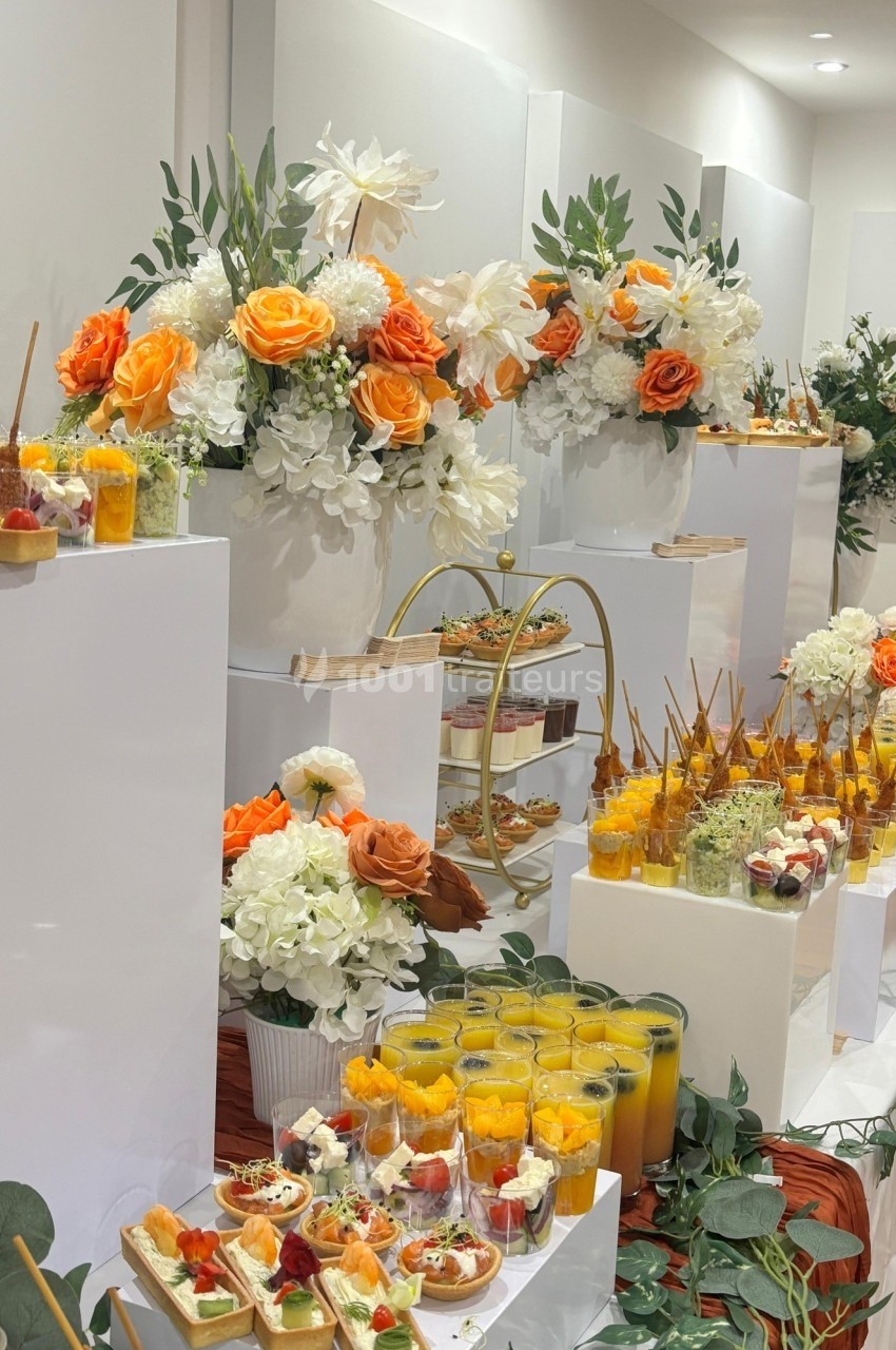 Buffet élégant avec fleurs orange et blanches, verrines, jus et assortiments de mets sur des présentoirs blancs.