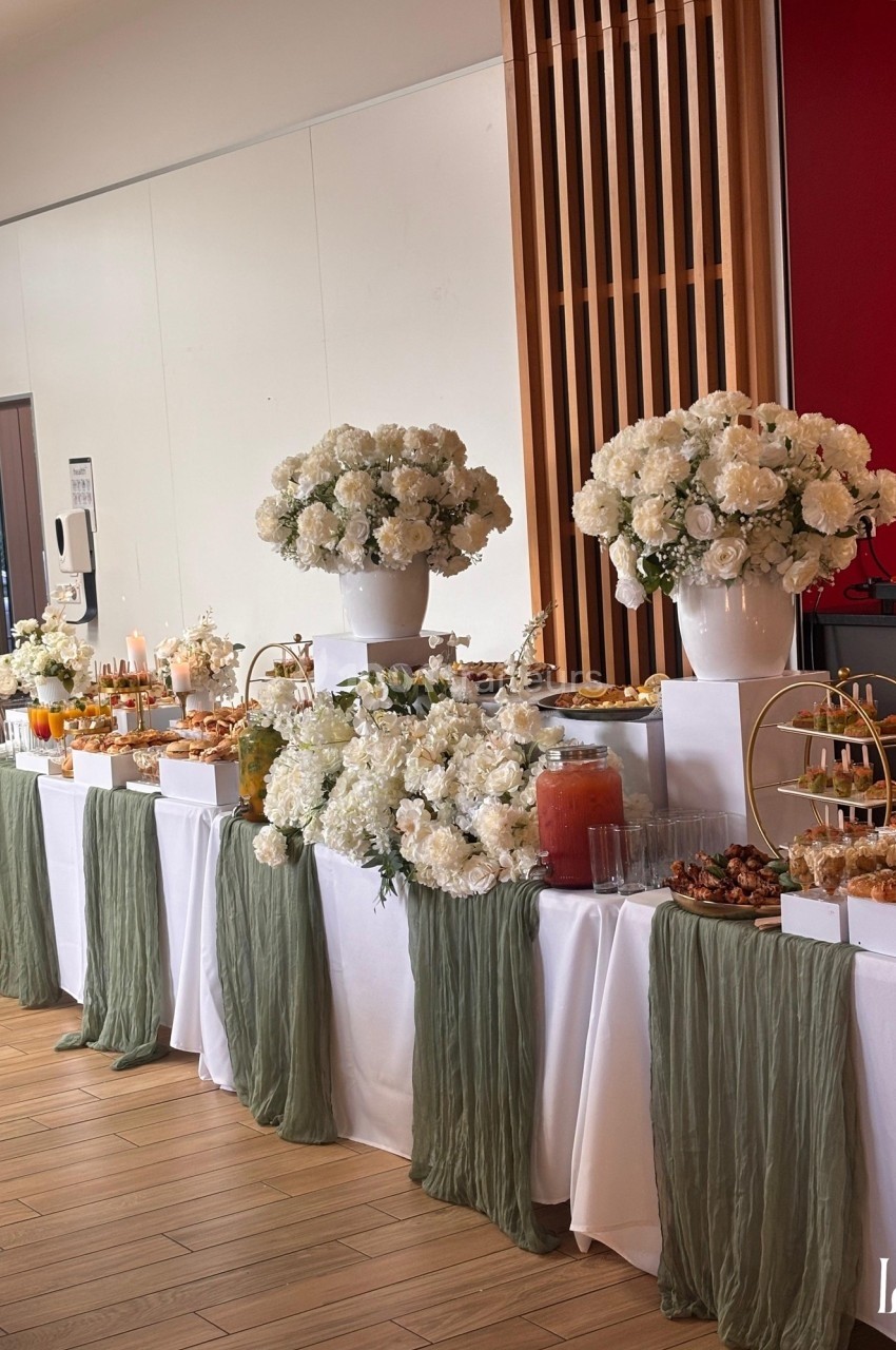 Table décorée pour un buffet avec fleurs blanches, nappes vertes, boissons et plats variés dans une salle lumineuse.
