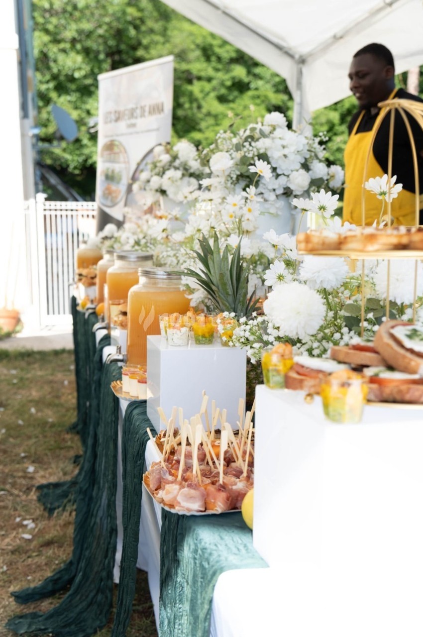 Buffet en plein air avec boissons, fleurs blanches, fruits et amuse-bouches disposés sur une table décorée.