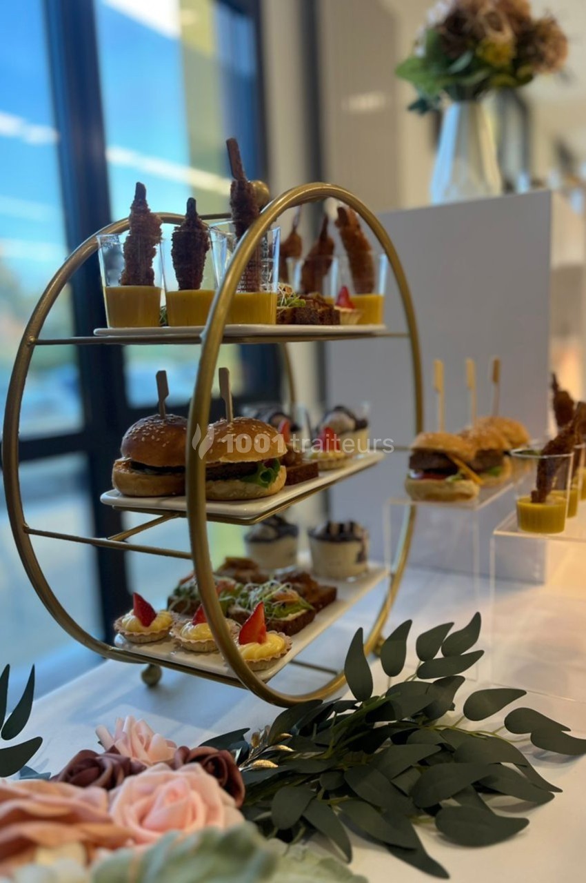 Présentoir doré avec des amuse-bouches variés, incluant mini-burgers, verrines et toasts, sur une table décorée.