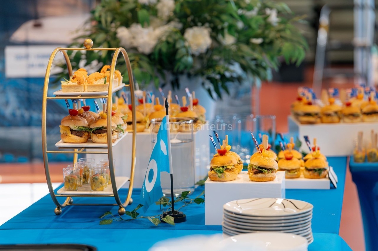 Buffet avec mini-burgers, verrines et décorations sur une table bleue, entourée de fleurs et vaisselle blanche.