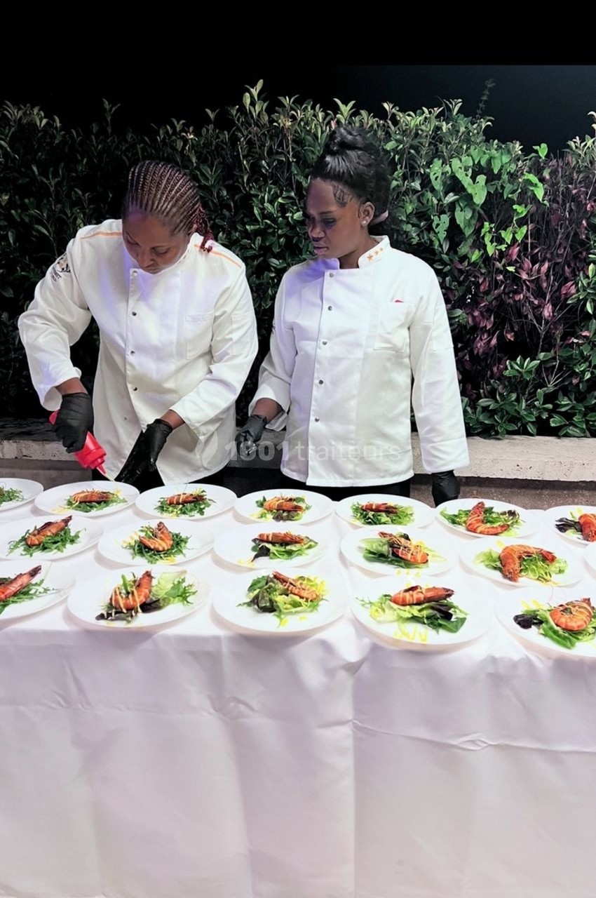 Deux chefs en tenue blanche dressent des assiettes garnies de légumes et de poisson sur une table blanche en extérieur.
