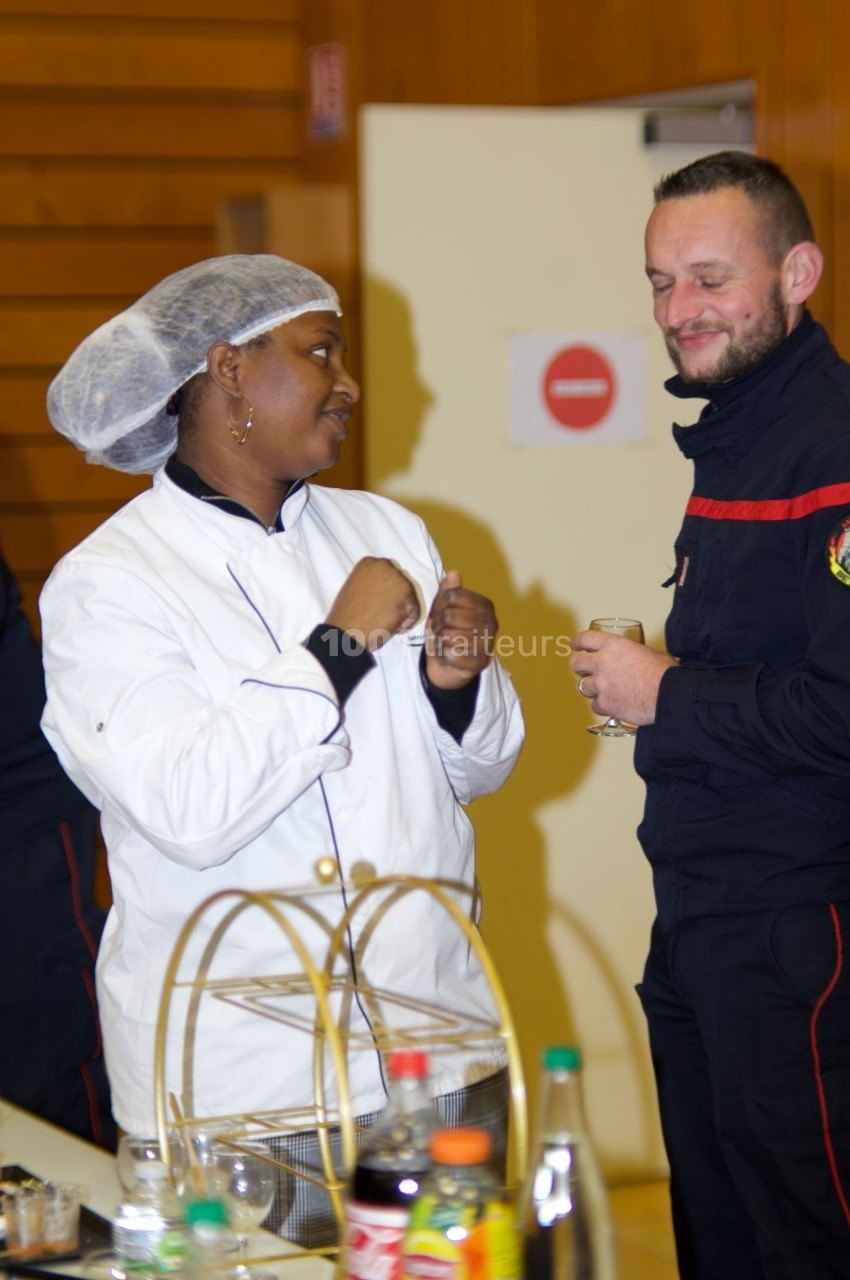 Une cuisinière échange avec un pompier dans une salle, près d'une table contenant des boissons et des aliments.