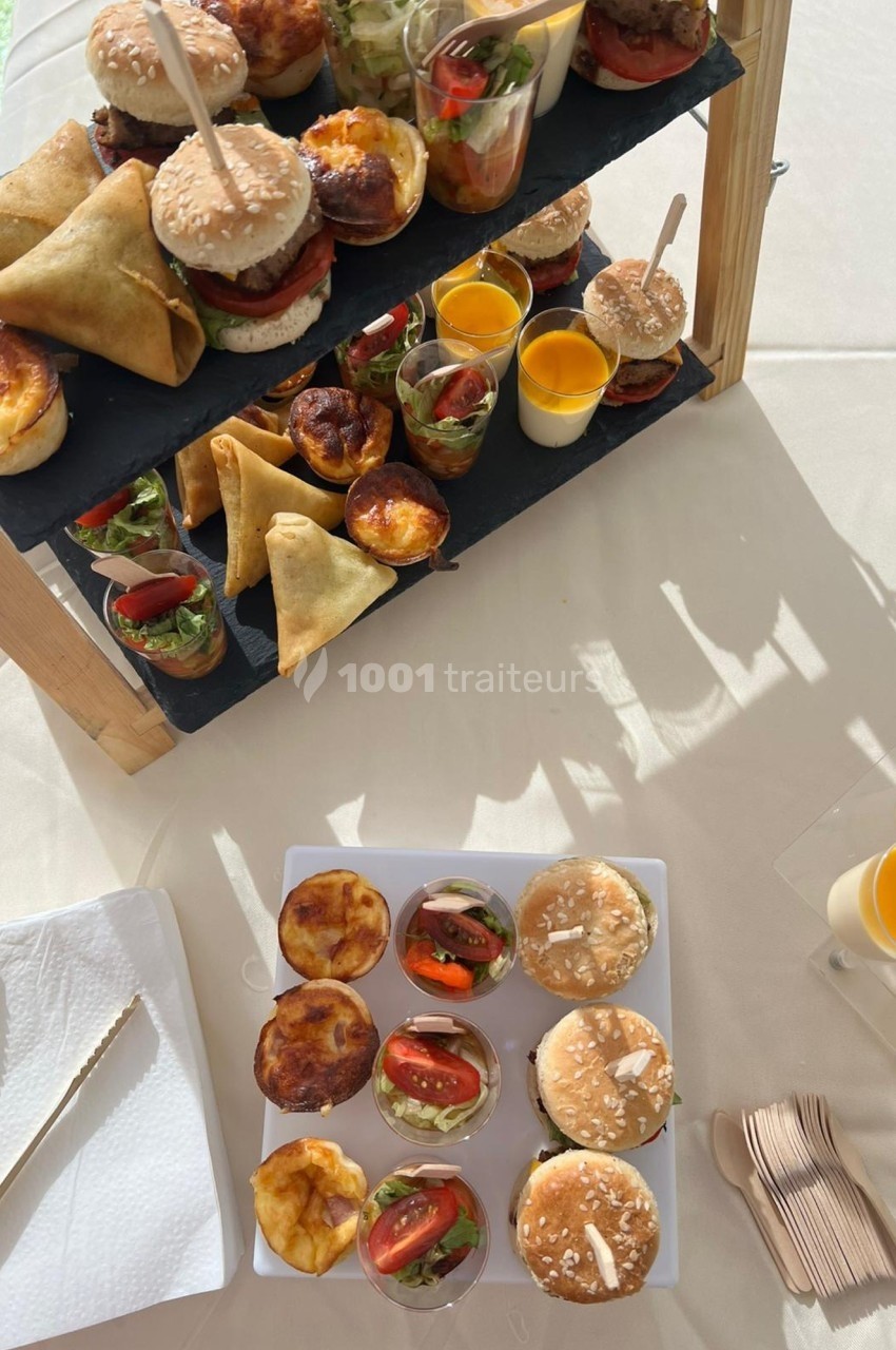 Plateau de mini-burgers, samoussas, quiches et verrines sucrées et salées disposés sur une table ensoleillée.