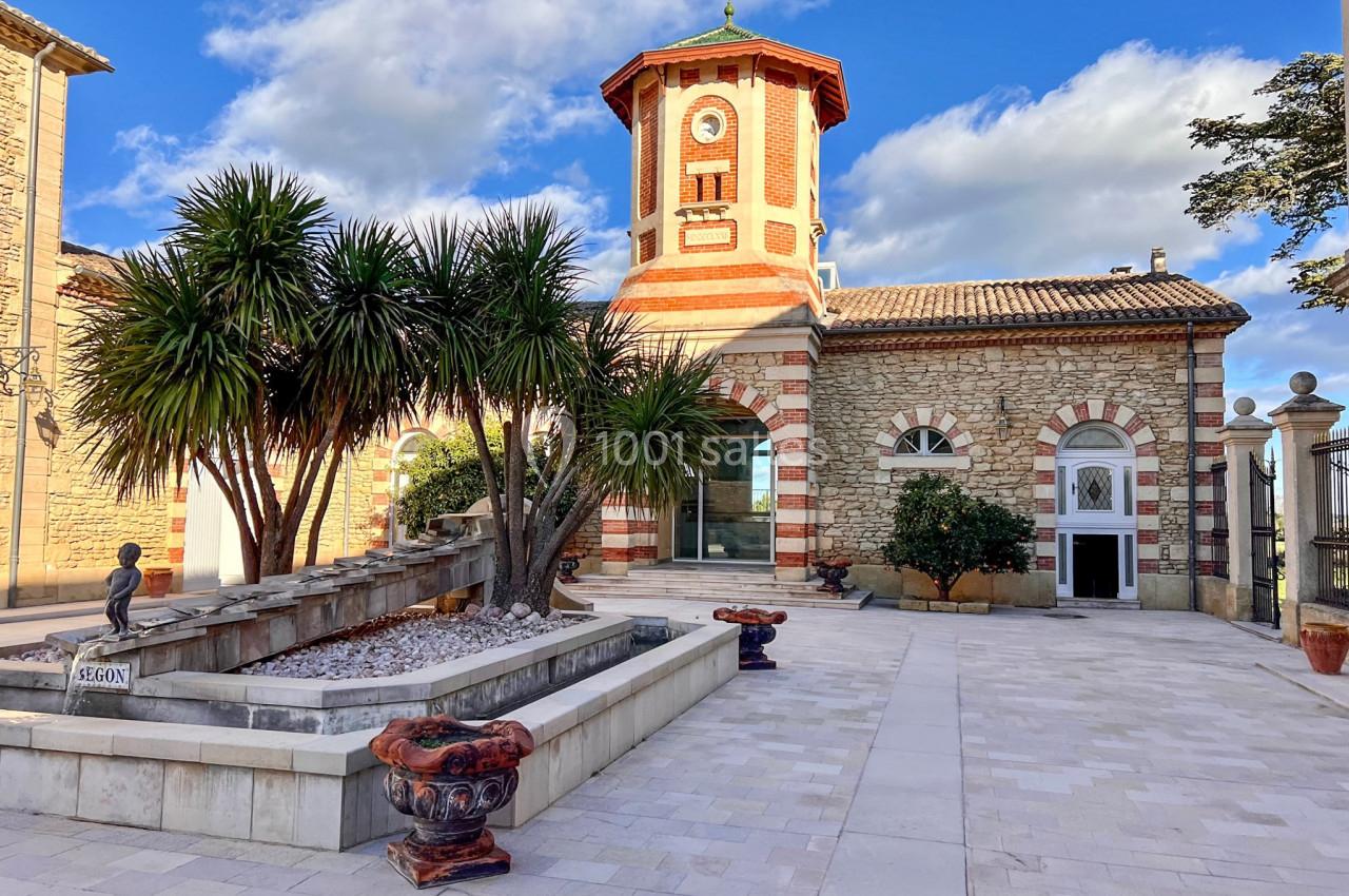 Bâtiment en pierre avec une tour octogonale, entouré de palmiers et d'une fontaine sur une place pavée.