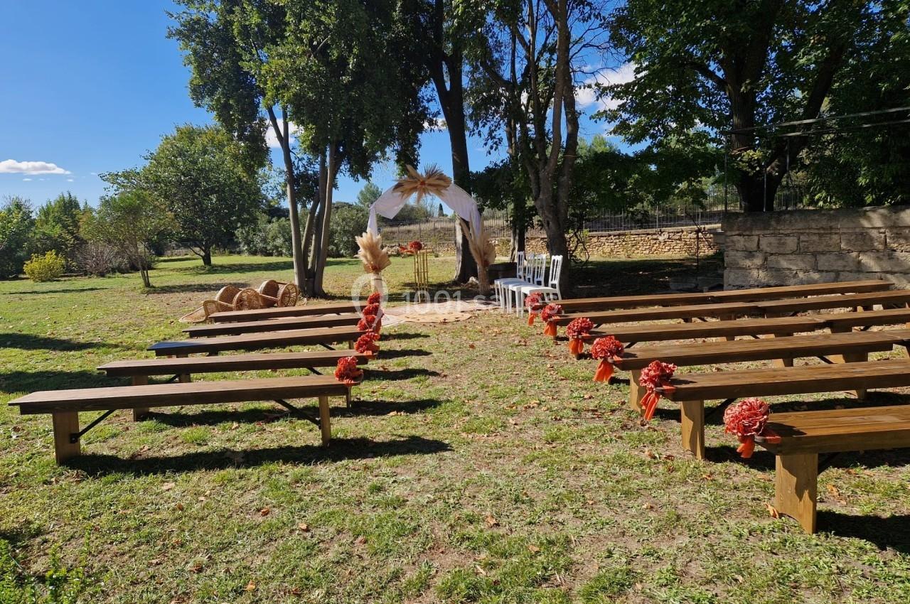 Allée centrale décorée de bancs en bois et fleurs rouges, menant à une arche de mariage dans un jardin ensoleillé.
