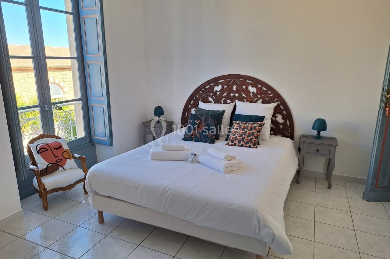 Chambre lumineuse avec lit double, tables de chevet, fauteuil et grande fenêtre donnant sur l'extérieur.