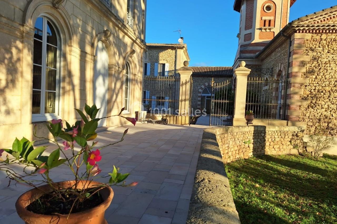 Façade en pierre d'un bâtiment ancien avec une tour, une terrasse ensoleillée et un pot de fleurs au premier plan.