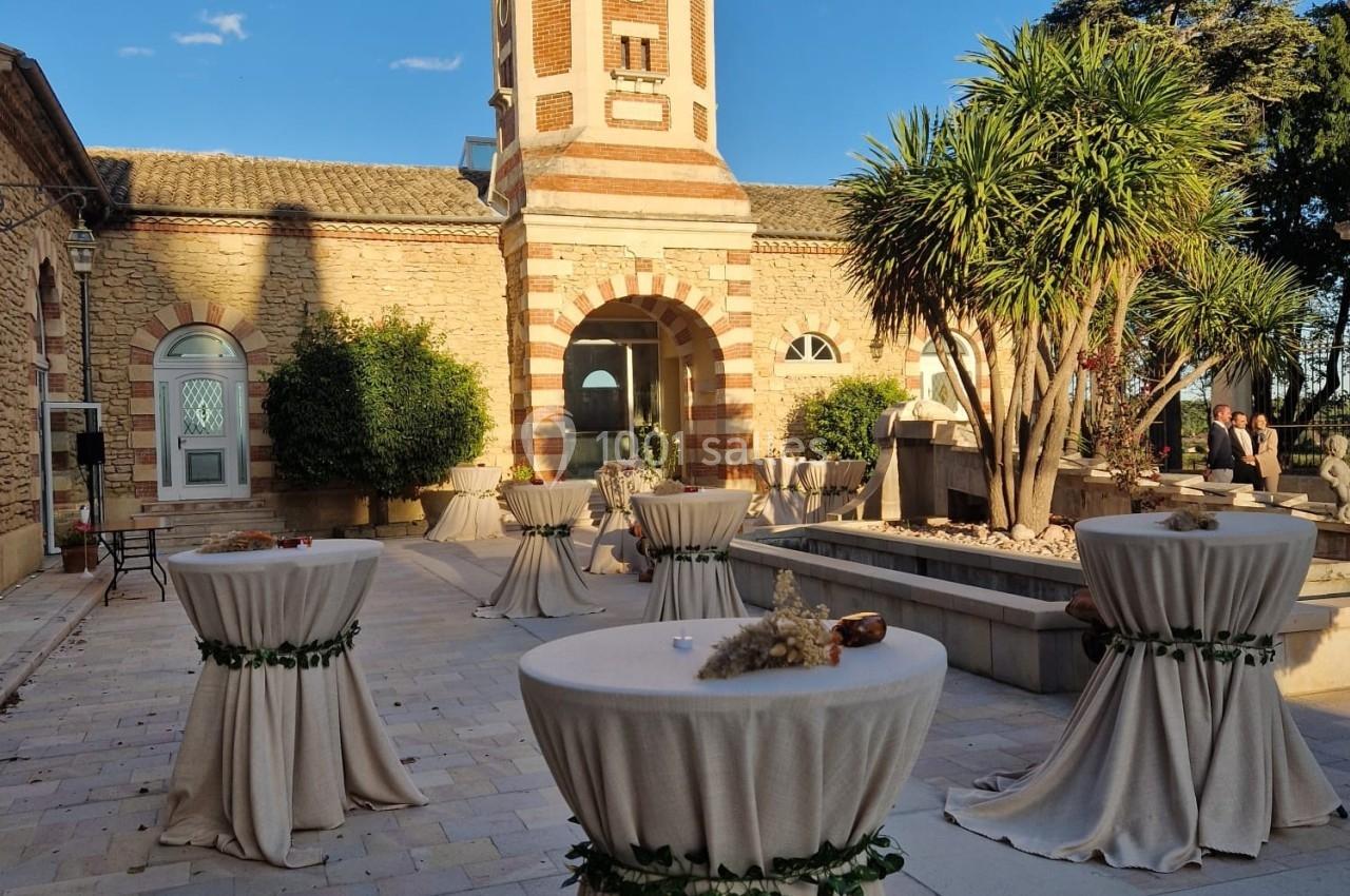 Cour extérieure avec tables hautes drapées de tissu beige, entourée de bâtiments en pierre et d'un clocher central.