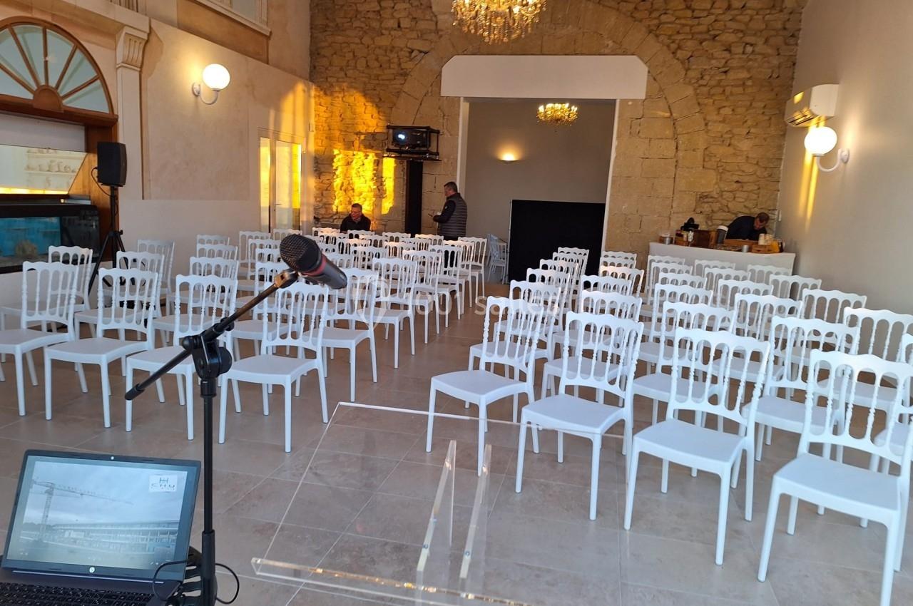 Salle de conférence lumineuse avec rangées de chaises blanches, un pupitre et un micro au premier plan.