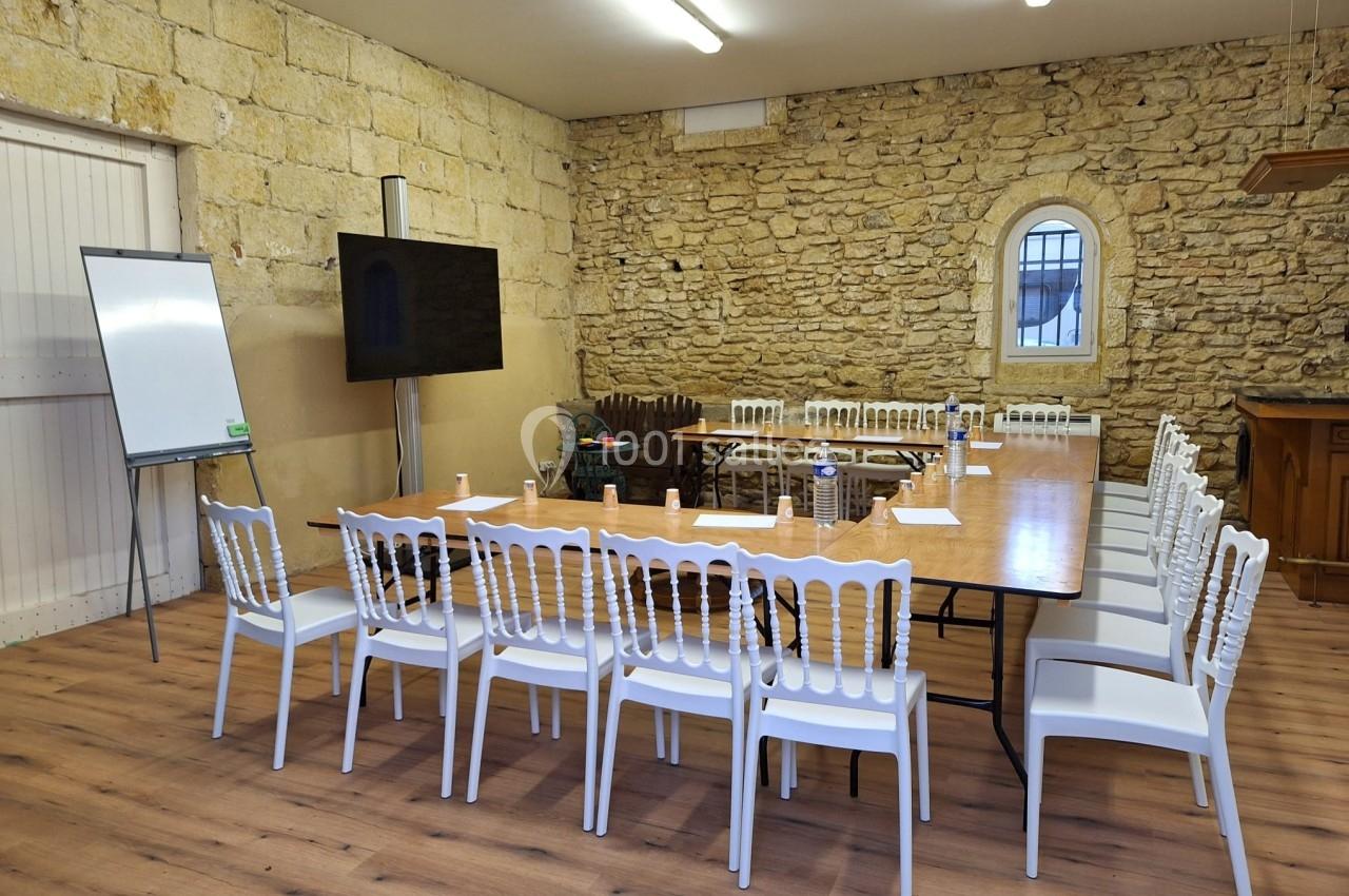 Salle de réunion avec murs en pierre, table rectangulaire entourée de chaises blanches, tableau blanc et écran.