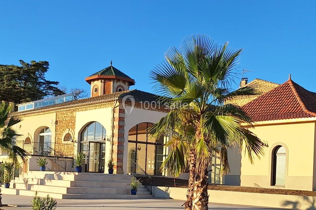 Bâtiment lumineux de style méditerranéen avec palmiers au premier plan et ciel bleu en arrière-plan.
