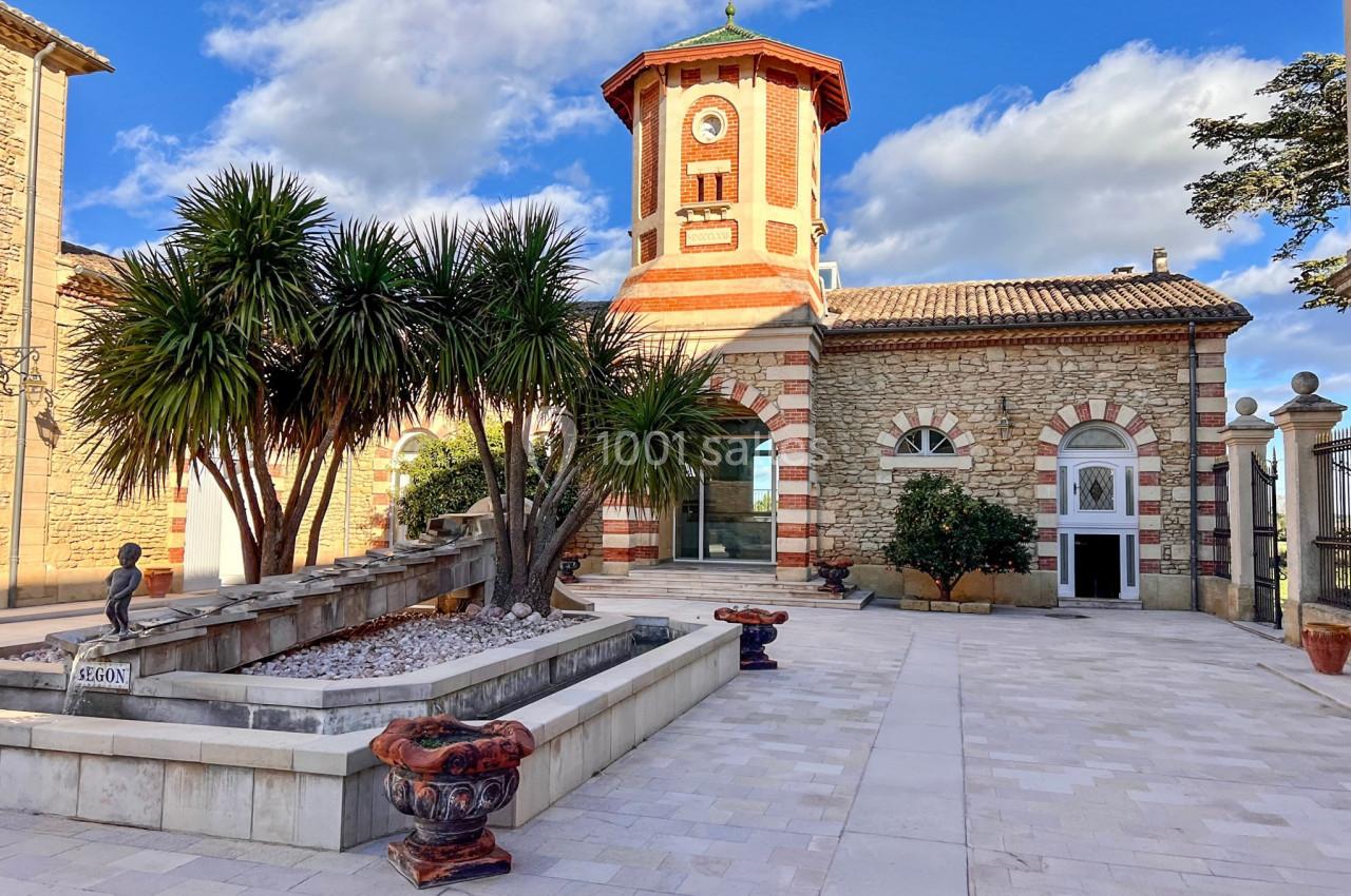 Bâtiment en pierre avec une tour octogonale, entouré de palmiers et d'une fontaine sur une place pavée.