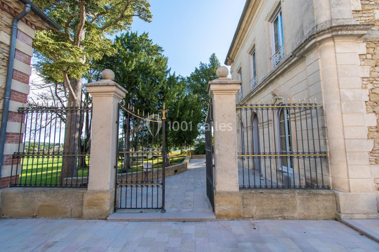 Portail en fer forgé ouvert, encadré par des piliers en pierre, donnant sur une allée bordée d'arbres et d'un bâtiment…