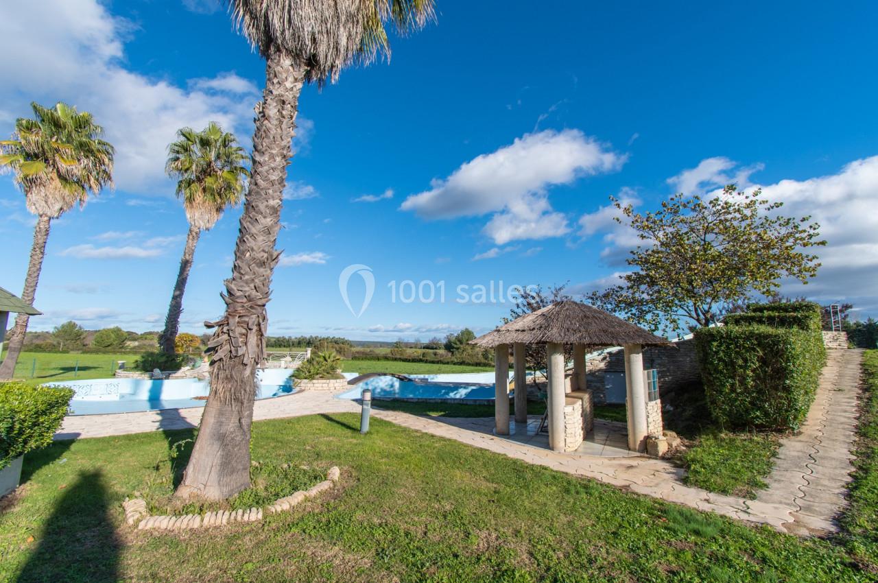 Jardin avec piscine, palmiers, pelouse et petit abri en chaume sous un ciel bleu parsemé de nuages.