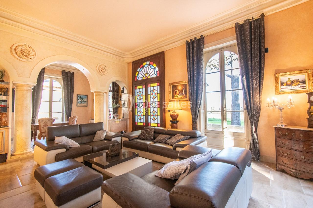 Salon lumineux avec canapés en cuir, vitraux colorés, grandes fenêtres, rideaux sombres et décorations classiques.