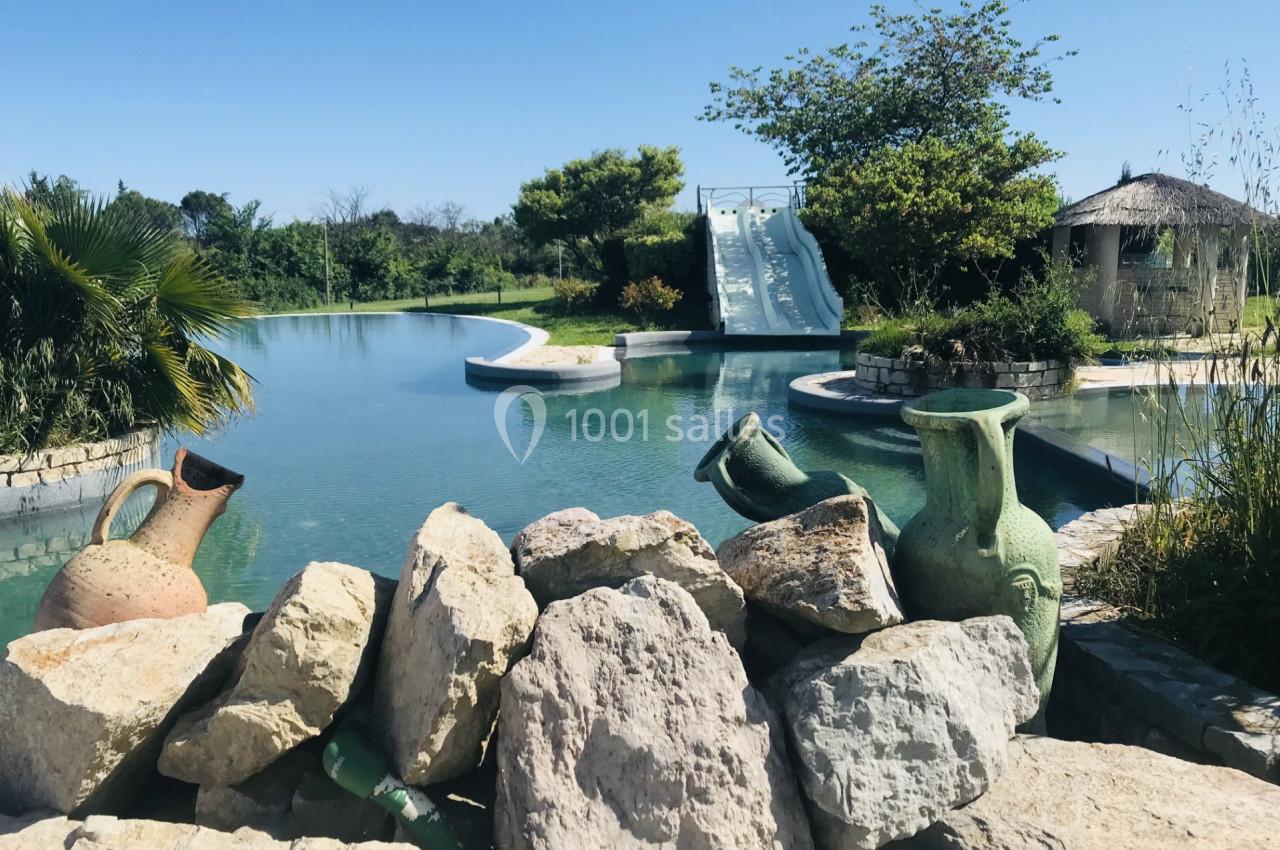 Vue d'un bassin avec toboggans aquatiques, entouré de végétation, de rochers et de jarres décoratives.