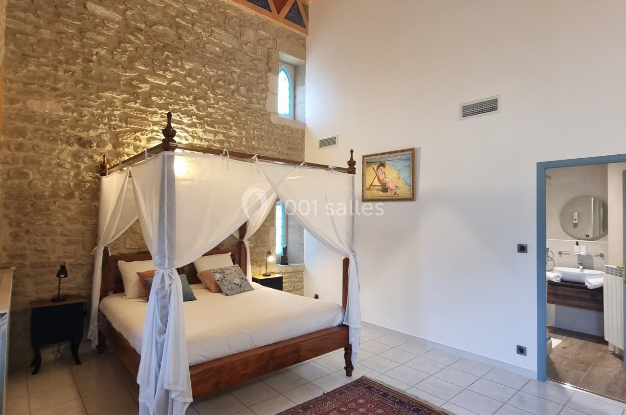 Chambre lumineuse avec lit à baldaquin, murs en pierre apparente, tapis coloré et plafond avec poutres en bois.