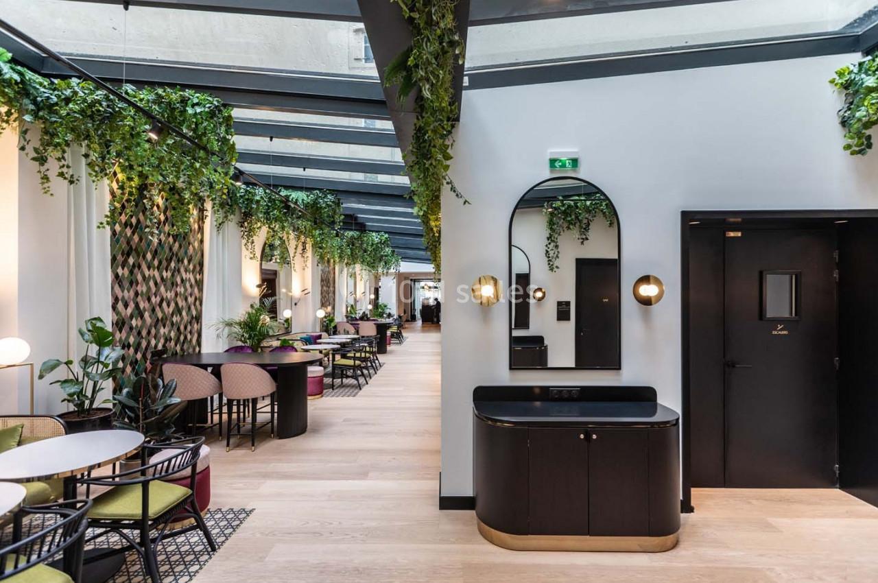 Salle lumineuse avec verrière, tables et chaises modernes, plantes suspendues et décoration élégante.