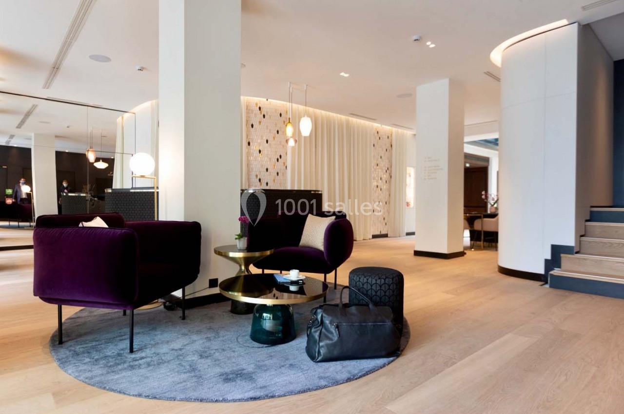 Espace salon moderne avec fauteuils violets, tables dorées, tapis rond gris et éclairage design dans un intérieur lumineux.