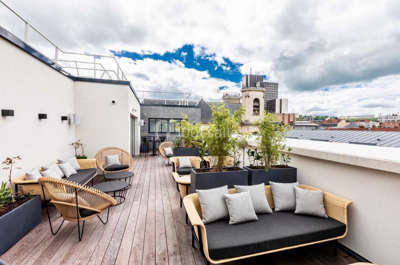 Terrasse en bois aménagée avec des canapés, des fauteuils et des plantes, offrant une vue sur des bâtiments urbains.