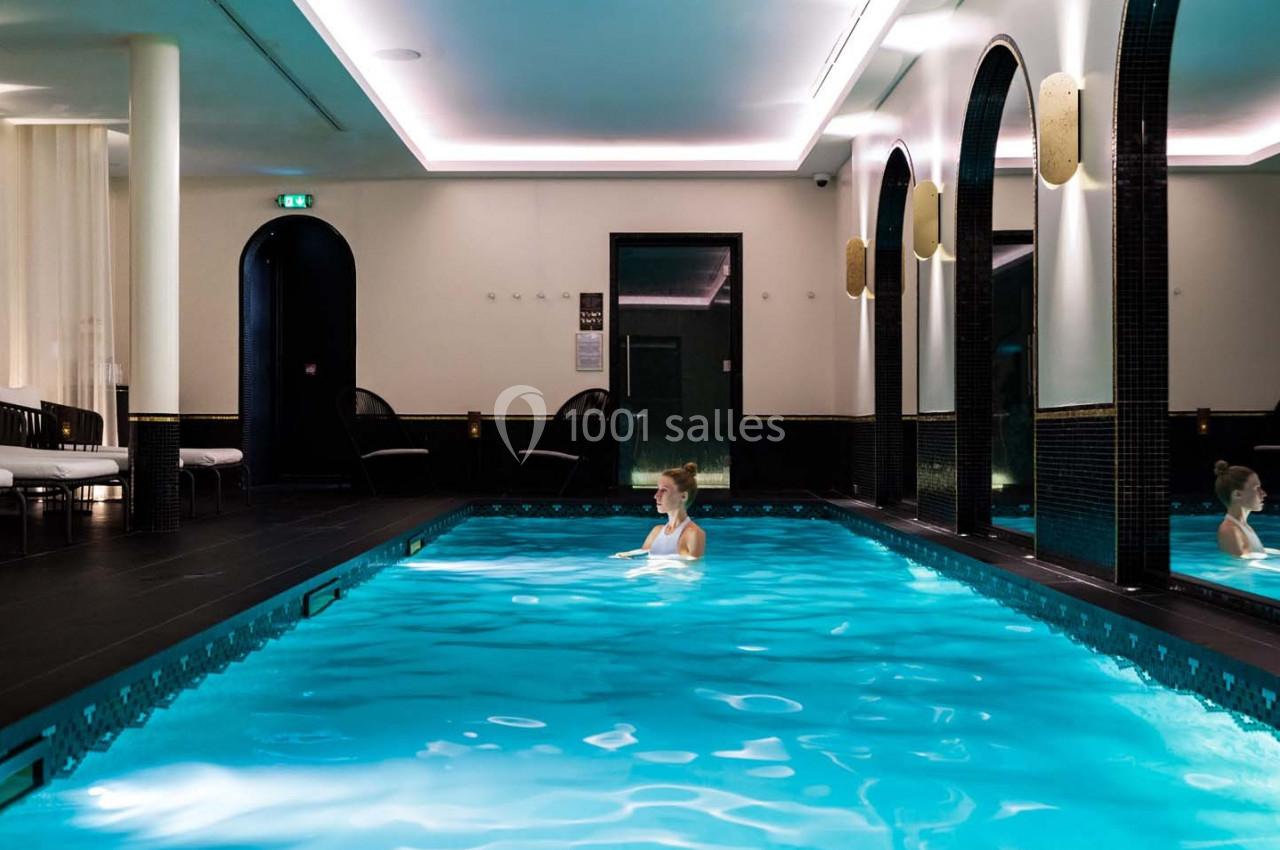 Femme seule dans une piscine intérieure éclairée, entourée de murs sombres avec des arches et des lumières tamisées.