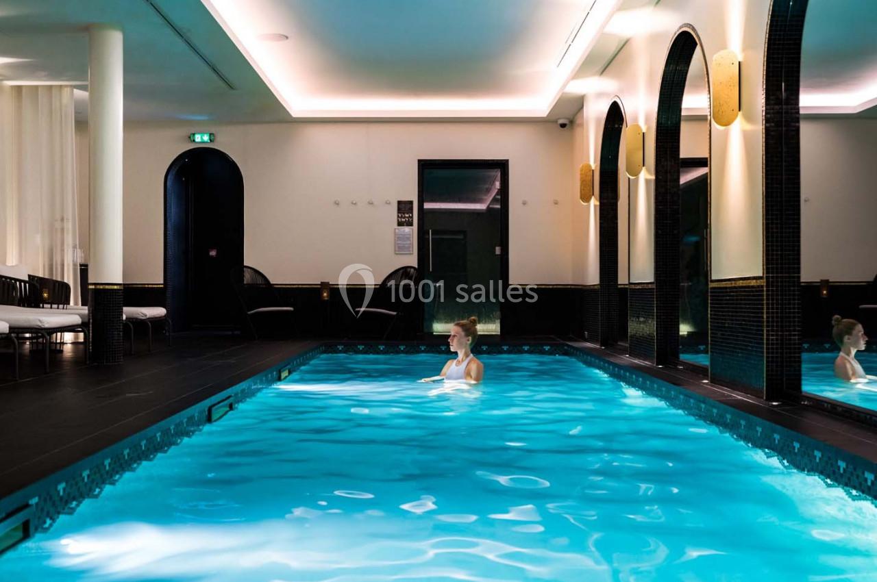 Femme seule nageant dans une piscine intérieure éclairée, entourée de murs sombres et d'arcs en miroir.