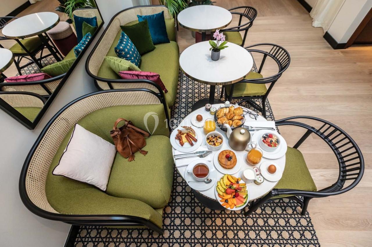 Table basse dressée pour un petit-déjeuner avec viennoiseries, fruits et boissons, entourée de fauteuils verts.